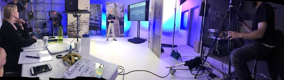 A professional film stage showing Dr. Shertz standing in front of high risk environment stage pieces and a television projecting slides while the crew watches with rapt attention